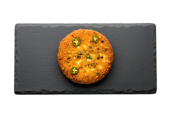 A golden-brown, spiced round pastry rests attractively on a dark gray slate plate, highlighted by vibrant green chili peppers.