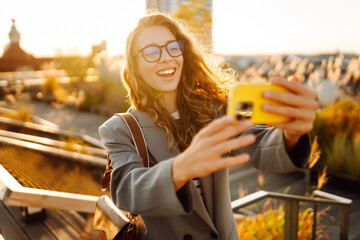 Portrait of a stylish woman wearing glasses and holding a phone, standing on a sunny rooftop. A beautiful woman takes a selfie on her phone, blogging outdoors. Blogging concept, technology. Lifestyle.