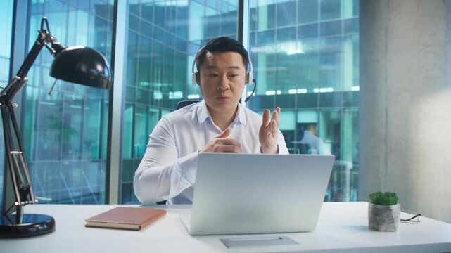 Engaged Asian man in headset sitting at desk speaking during video call in glass office. Communicating with client or presenting service. Answering questions or offering consultation online.