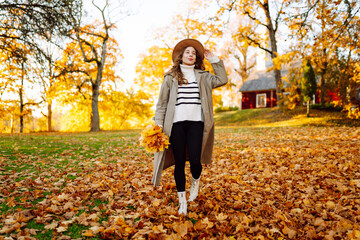 A happy woman is having fun in an autumn park at sunset. A beautiful woman in casual clothes throws leaves into the air on a sunny day. Concept of a walk, relaxation.