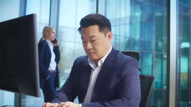 Focused Asian male in navy suit typing on keyboard in corporate office. Preparing report or answering email. Blonde woman in background speaking on phone while standing near window.