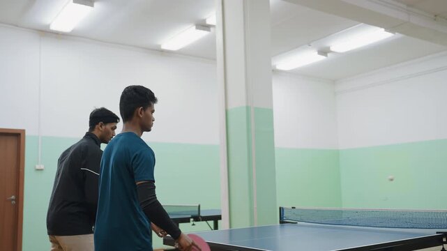 Young contender in blue shirt faces elderly coach during indoor table tennis match, both focused on ball crossing net, training hall with green and white walls and bright lighting