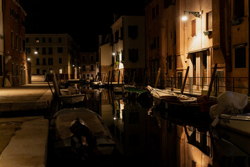 Venetian canal at night with moored boats and glowing reflections, warm streetlamps illuminating...