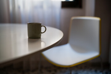 Serene morning composition with green mug on bright white tabletop near yellow-edged chair, warm daylight and gentle shadows highlighting simplicity and comfort in the room