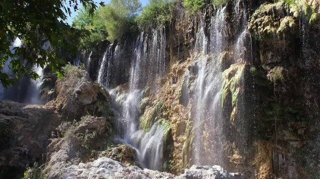 Multi-level waterfall cascades down a rugged rock face surrounded by greenery. Sunlight sparkles through mist. Natural wonder, eco tourism and fresh environment concept. 