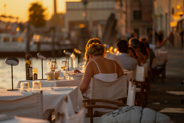 People enjoying outdoor evening dinner in Venice, Italy by the sea at sunset, warm golden light reflecting on tables with wine and food, creating romantic holiday mood of coastal summer evenings