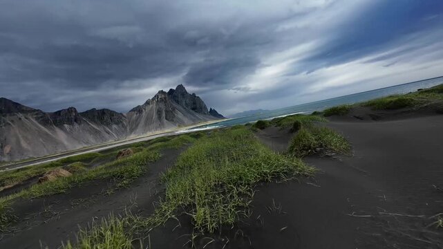 An immersive FPV drone flight starts over Stokksnes' grass dunes under a dramatic sky, flying low above the black sand beach with waves towards Vestrahorn mountain.