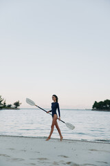 A solo female paddler walks along a tranquil beach, carrying a paddle, wearing a dark swimsuit as calm water and a pale sky create a peaceful coastal moment.