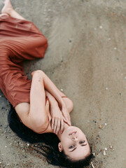 A young woman lies on sandy ground in a rust colored dress, arms folded near her chest, head tilted, soft light, intimate mood, delicate portrait of calm.