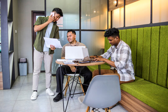 Three Men Working on Project in Modern Office, With One on Phone Call While Others Focus on Laptop - Powered by Adobe