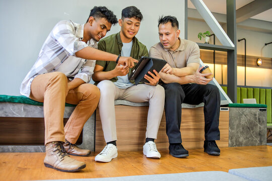 Diverse Young Men Sitting Indoors, Engrossed in Discussion While Using Digital Tablet - Powered by Adobe