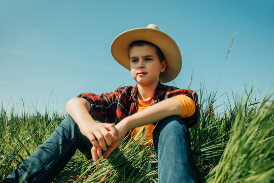 Cowboy kid in hat sits on grass daydreaming on a sunny day outdoors