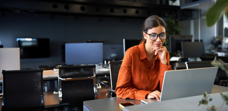 Smiling busy financial specialist latin hispanic businesswoman working on laptop pc, watching educational video in office. European mature woman using computer for finance data analysing work online.