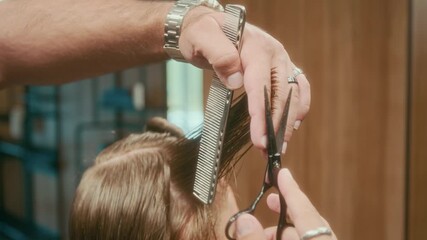 Close-up of hands of unrecognizable professional male hairstylist cutting red hair of regular customer using fine comb and scissors, pulling strand upwards and trimming ends