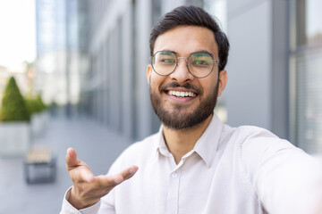 Young indian man in glasses smiling at camera while vlogging outdoors in a modern city, gesturing confidently as a content creator and entrepreneur sharing ideas on video