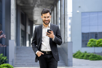 Young businessman walking outside modern office building, smiling, checking social media, messaging on mobile phone, using corporate app, communicating with colleagues
