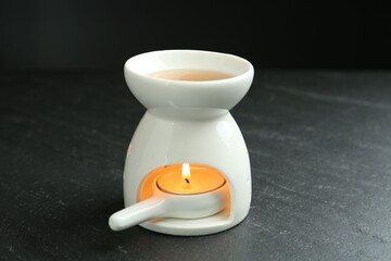 Aroma lamp with burning candle on dark textured table against black background, closeup