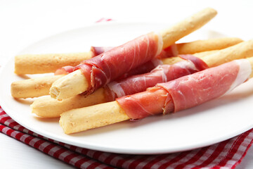Delicious grissini sticks with prosciutto on white wooden table, closeup