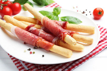 Delicious grissini sticks with prosciutto, peppercorns and basil on white wooden table, closeup