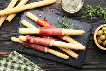 Delicious grissini sticks with prosciutto, peppercorns, green olives and rosemary on wooden table, flat lay