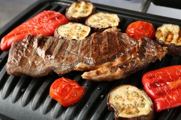 Electric grill with beef steaks and vegetables on grey table, closeup