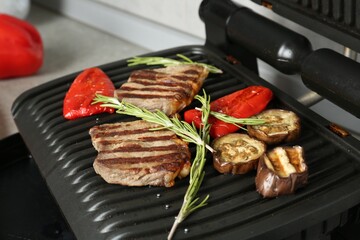 Open electric grill with beef steaks, rosemary and vegetables on table, closeup