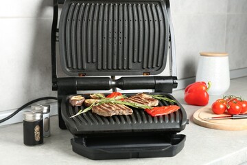 Open electric grill with beef steaks, rosemary, vegetables and spices on light grey table, closeup