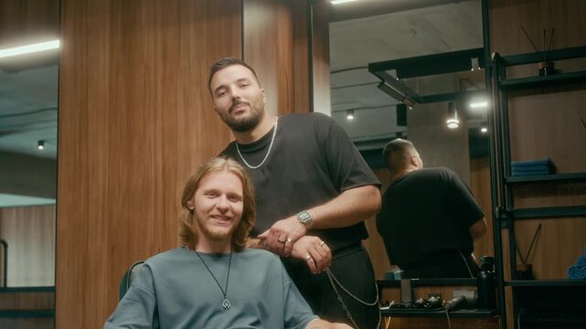 Medium portrait of handsome young Caucasian barber in black tshirt tousling male customers hair while giving styling advice after haircut, bot looking at camera and smiling