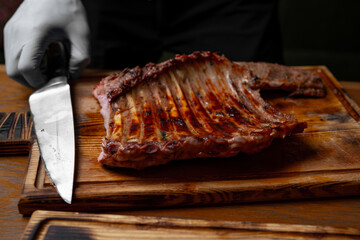 Juicy grilled pork ribs on wooden cutting board with chef’s knife and gloved hand in restaurant kitchen, close up of roasted barbecue meat preparation