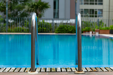 Swimming pool with stair and wooden deck in front with blurred green background. Outdoors on sunny day. Space for text. Poolside and outdoor spa vacation day.Grab bars ladder in the blue swimming pool