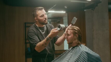 Medium shot of professional young Caucasian barber styling male clients long red hair with hairdryer and brush after giving haircut, with confident, skillful movements, while working at trendy salon