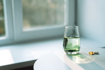 Glass of water with chlorophyll partially dissolved, soft green plumes. Capsules and softgels nearby. Horizontal wellness scene.