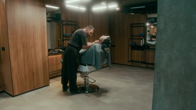 Wide shot of young Caucasian barber in black tshirt and pants styling and shaping beard for mature male client, relaxing in reclining chair, then stepping away to check result