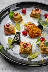Elegant gourmet appetizer plate with assorted canapés topped with pâté and berry sauce, caramelized fruit, microgreens, fresh raspberries, blackberries, and grapes arranged on a black ceramic plate