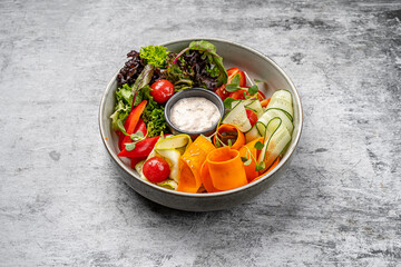 Healthy fresh vegetable salad with cucumber ribbons, carrot curls, lettuce, cherry tomatoes, bell pepper, microgreens, and creamy dipping sauce served in a modern bowl