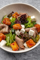 Fresh salad with mixed greens, roasted meat slices, feta cheese cubes, and crispy glazed croutons served in a ceramic bowl on gray table background