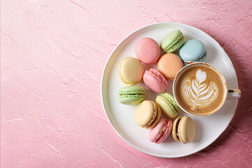 Multicolored French macarons on a plate, next to a cup of cappuccino with a pattern on a light pink vintage texture background. Top view.