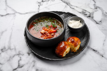 Traditional Ukrainian borscht soup with beetroot, cabbage, pork, fresh herbs, served in black ceramic bowl with sour cream and homemade golden buns on marble background