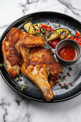 Roasted chicken leg quarter with crispy golden skin served on black plate with grilled vegetables, chili dipping sauce, pink peppercorns and fresh microgreens on marble background