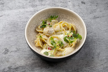 Traditional dumplings with meat filling served in a bowl with creamy sauce, microgreens and pink peppercorns on gray background