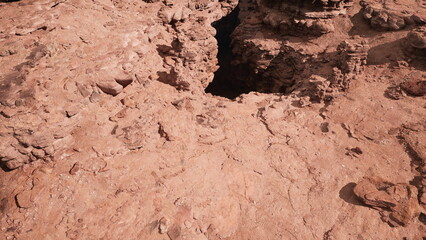 Rugged terrain showcases an intricate rock structure with a significant crevice, bathed in sunlight. Dusty earth and weathered rocks tell stories of natures forces at play in this barren environment.