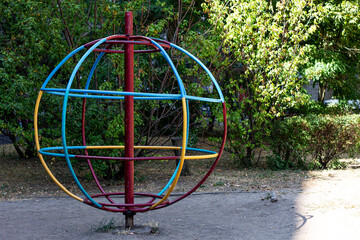 A vibrant, colorful spinning playground orb stands in a park, its bright red, blue, and yellow rings contrasting with the green bushes and dappled sunlight.

