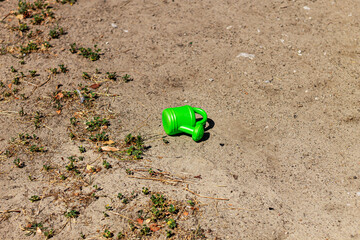 A bright green watering can lies on the parched ground, its vibrant color creating a sharp contrast against the dry earth. The image evokes a quiet sense of summer nostalgia.

