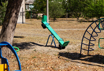 A lonely seesaw sits on a playground in a sunlit park, surrounded by dry grass and fallen leaves. The image captures a quiet, nostalgic moment of childhood and summer.


