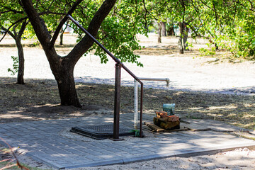 An old water pump stands in a peaceful, sunlit park. The scene captures a quiet moment of rural life, with green trees and natural light highlighting the weathered texture of the pump.

