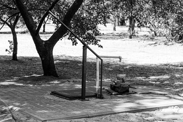 An old manual water pump stands on a paved brick square under the shade of trees on a sunny day. The black and white tones add a sense of timelessness.

