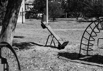 An empty seesaw and playground equipment on a sunny day, with long shadows cast across the ground, captured in a black-and-white image.

