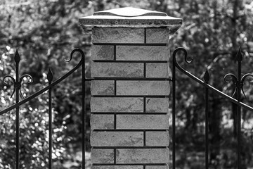 A detailed, close-up, monochrome shot of a brick pillar and an ornate wrought iron fence against a blurred background of trees, highlighting textures.

