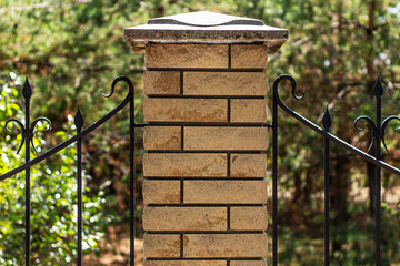 A detailed, close-up shot of a brick pillar with a decorative wrought iron fence on a sunny day, with soft-focus green foliage in the background.

