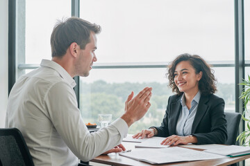 Fototapeta premium man discussing business with smiling Middle Eastern woman at office desk, both sitting across from each other with documents and glass of water visible